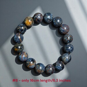 Puede incluir: Una pulsera circular compuesta por diecis&eacute;is cuentas esf&eacute;ricas pulidas con patrones de remolinos en tonos azul profundo, gris y marr&oacute;n dorado. La pulsera est&aacute; marcada como #8 y tiene una longitud total de 16 cm, seg&uacute;n indica el texto.