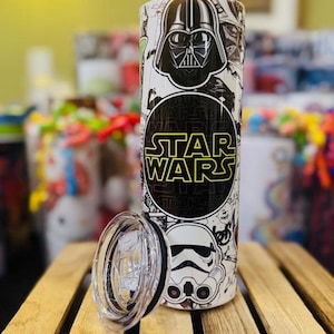 Könnte beinhalten: Ein weißer Star Wars-Becher mit einem schwarz-weißen Design mit Darth Vader und einem Sturmtruppler. Der Becher enthält die Worte "STAR WARS" in einem gelb-schwarzen Kreis. Der transparente Deckel liegt daneben.