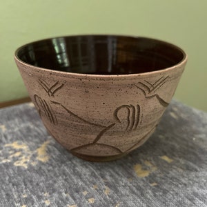 May include: A handmade ceramic bowl with a light brown exterior and a dark brown interior. The bowl features etched geometric patterns and a speckled texture. The bowl is displayed on a gray surface.