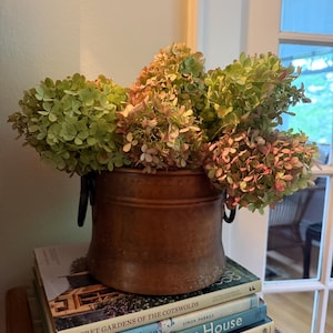 May include: A copper bucket with two side handles containing a large bouquet of dried hydrangea flowers in shades of green and muted pink. The bucket rests on a stack of four hardcover books titled Secret Gardens of the Cotswolds and Summer House.