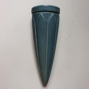May include: A cone-shaped wall vase with a matte, dusty blue finish. The ceramic vessel features a series of vertical, pointed petal-like ridges that extend from the circular opening down to the tapered, rounded point at the bottom of the object.