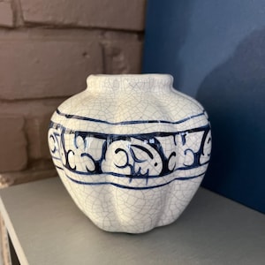 May include: A small, round ceramic vase with a crackle glaze finish. The vase features a band of blue decorative patterns around the middle. The vase is sitting on a gray shelf.