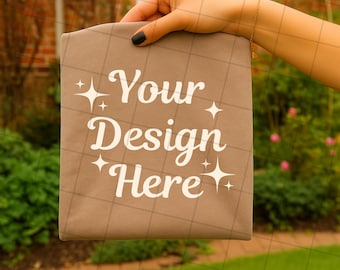 May include: A tan fabric item with the words "Your Design Here" in white script. The item is held by a hand with black nail polish, against a blurred outdoor background of greenery and a brick building.