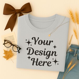 May include: A light gray sweatshirt with the text "Your Design Here" in black script. The image includes a brown bow, eyeglasses, a gold necklace, a gold pen, a leaf, and a pair of blue jeans.