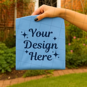 May include: A light blue fabric item with the text "Your Design Here" in dark blue script. The text is surrounded by starburst graphics. The fabric has a grid pattern and is held up by a hand.