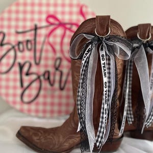 Puede incluir: Botas de vaquero de cuero marrón con cintas decorativas en blanco y negro. Las cintas tienen el texto "GO TEAM!". Un letrero rosa y blanco a cuadros que dice "Boot Bar" está en el fondo.