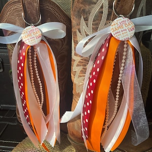 May include: Two brown leather cowboy boots with decorative keychains. Each keychain has a button with the text "GAME DAY" and red, orange, and white ribbons, along with a string of pearls. The boots are on a burlap surface.