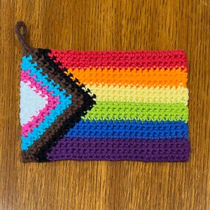 Peut inclure: Un objet crocheté fait à la main représentant un drapeau arc-en-ciel avec une boucle marron. Le drapeau comprend les couleurs arc-en-ciel traditionnelles, ainsi que du noir, du marron, du bleu clair, du rose et du blanc. Il s'agit probablement d'un dessous de plat ou d'une petite pièce décorative.