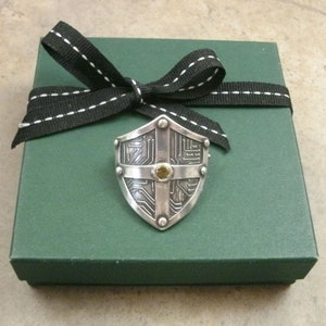 Pu&ograve; includere: Un ciondolo a forma di scudo in argento con un design a circuito stampato e una piccola pietra preziosa gialla al centro. Il ciondolo &egrave; in una scatola regalo verde con un nastro nero.