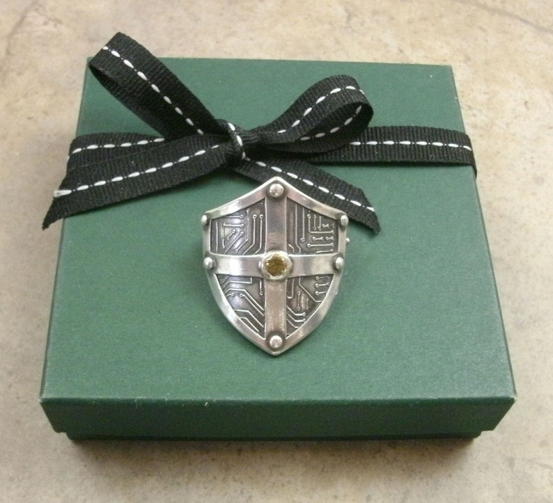 Pu&ograve; includere: Un ciondolo a forma di scudo in argento con un design a circuito stampato e una piccola pietra preziosa gialla al centro. Il ciondolo &egrave; in una scatola regalo verde con un nastro nero.