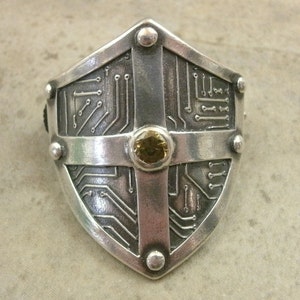 Pu&ograve; includere: Un anello a forma di scudo in argento con un design a circuito stampato e una pietra preziosa gialla al centro.
