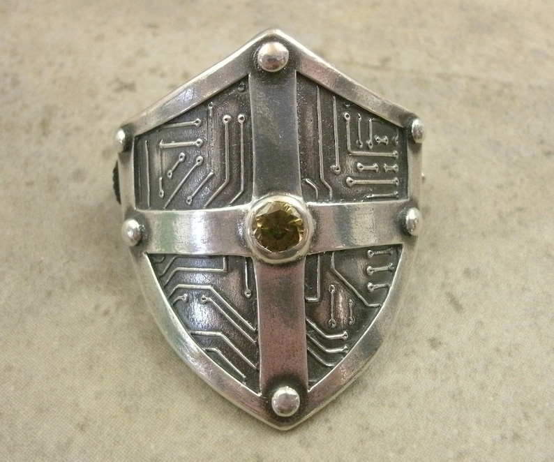 Pu&ograve; includere: Un anello a forma di scudo in argento con un design a circuito stampato e una pietra preziosa gialla al centro.