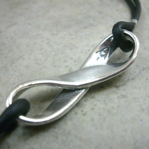 May include: A silver infinity symbol pendant on a black leather cord. The pendant is made of a twisted metal design and has a small mark on the side.
