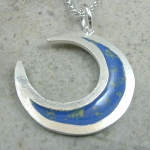 May include: A silver crescent moon pendant with a blue and green speckled enamel inlay.