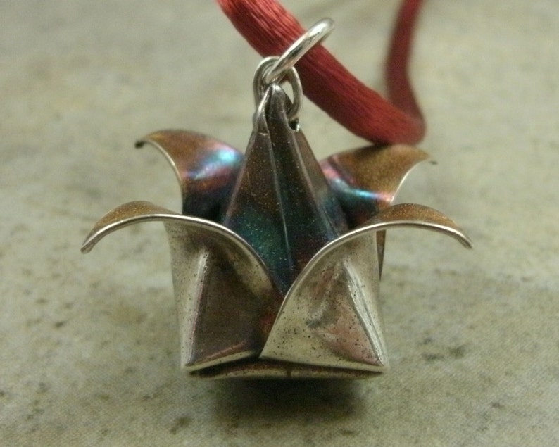 May include: A silver origami flower pendant with a red cord. The flower has a metallic sheen and is folded into a delicate shape.