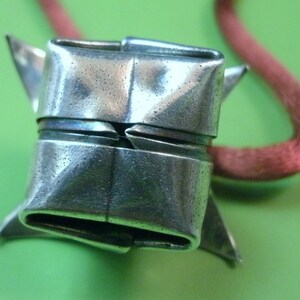 May include: A silver origami-style pendant with a folded design, hanging from a red cord.