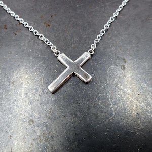 以下が含まれることがあります： 繊細なチェーンにシルバーのクロスチャームがついたネックレス。クロスはシンプルでミニマルなデザインで、わずかにテクスチャ加工が施されています。