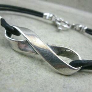 May include: A silver infinity symbol charm bracelet with a black leather strap. The charm is made of a smooth, polished metal and has a simple, elegant design.