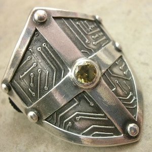 Pu&ograve; includere: Un anello d'argento a forma di scudo, con un design a circuito stampato e una pietra preziosa gialla al centro.
