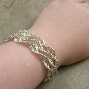 Puede incluir: Un brazalete de cadena de plata con un diseño tejido. El brazalete se lleva en la muñeca.