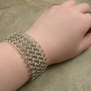 May include: A silver chainmail bracelet with a woven pattern. The bracelet is worn on a person's wrist.