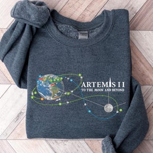 May include: Dark gray sweatshirt with the text "ARTEMIS II TO THE MOON AND BEYOND" and a graphic of Earth, the moon, and connecting lines. The sweatshirt is folded on a light brown and white wooden surface.