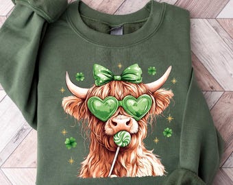 May include: Military green sweatshirt featuring a Highland cow wearing heart-shaped sunglasses and a bow, holding a lollipop. The cow has long, shaggy brown fur and white horns. The sweatshirt has the text "MILITARY GREEN" at the top.