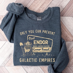 May include: A dark grey sweatshirt featuring the text "ONLY YOU CAN PREVENT Visit ENDOR Hiking-Camping-Feasting Lunar Forest GALACTIC EMPIRES" in gold. The design includes a cartoon image of a person in a ranger hat.