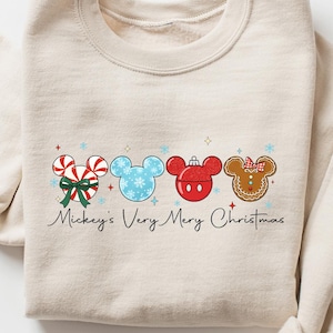 May include: Beige sweatshirt featuring a festive graphic with Mickey Mouse-themed Christmas ornaments. The design includes candy canes, snowflakes, and gingerbread, with the text "Mickey's Very Merry Christmas" in a script font. The sweatshirt is laid flat.