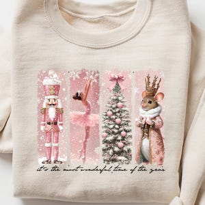 May include: Cream-coloured sweatshirt with a festive design. The design includes a nutcracker, ballerina, Christmas tree, and mouse in pink and white. The text "It's the most wonderful time of the year" is printed below the design.