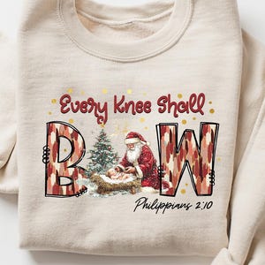 May include: Beige sweatshirt with the text "Every Knee Shall Bow" and the letters B, A, and W in a red and brown pattern. The design includes an illustration of Santa Claus and a Christmas tree. The text "Philippians 2:10" is also present.