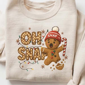 K&ouml;nnte beinhalten: Beiges Sweatshirt mit dem Schriftzug "OH SNAP" in Lebkuchen-Schrift. Ein Lebkuchenmann mit roter M&uuml;tze und Zuckerstange ist abgebildet. Das Design umfasst Lichterketten und Sterne, ideal f&uuml;r die Weihnachtszeit.