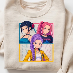 May include: Beige crewneck sweatshirt featuring a colorful anime-style graphic of three women. The graphic includes the word "Venom" on one woman's shirt. The design is framed by yellow and blue rectangles.