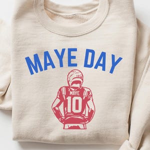 K&ouml;nnte beinhalten: Beiges Sweatshirt mit dem Text "MAYE DAY" in Blau und einer rot-wei&szlig;en Footballspieler-Grafik. Die Trikotnummer des Spielers ist 10. Das Sweatshirt hat lange &Auml;rmel.