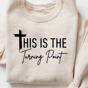 Peut inclure: Un sweat-shirt beige avec l'inscription noire "THIS IS THE Turning Point". Le design comprend une croix au-dessus des mots. Le sweat-shirt est plié, les manches retroussées.