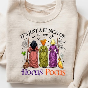 Peut inclure: Sweat-shirt beige avec un motif de trois sorcières et le texte "It's Just a Bunch of Hocus Pocus, Est. 1693". Les sorcières portent des robes roses, vertes et violettes, tenant des balais. Vêtement sur le thème d'Halloween.