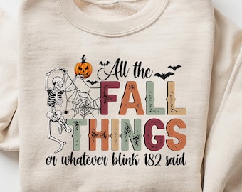 May include: Beige sweatshirt with the text "All the FALL THINGS or whatever blink 182 said." The design includes a skeleton, pumpkin, bats, and spiderwebs. The sweatshirt is folded with the sleeves rolled up.