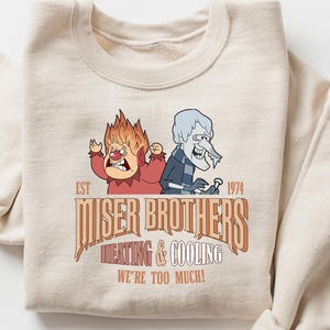 May include: Beige sweatshirt featuring a cartoon graphic of two characters, one fiery red and the other icy blue, with the text "Miser Brothers Heating & Cooling We're Too Much!" in brown and orange. The sweatshirt has rolled-up sleeves.
