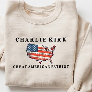 May include: Beige sweatshirt with the text "CHARLIE KIRK" above a graphic of the United States flag in the shape of the country. Below the graphic is the text "GREAT AMERICAN PATRIOT".