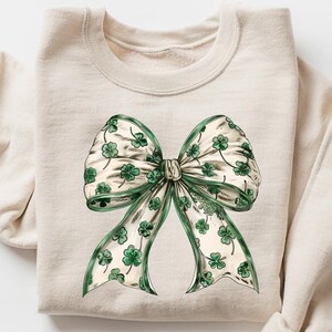 May include: A cream-colored sweatshirt featuring a large bow graphic. The bow is detailed with green shamrocks on a white background, with green trim. The sweatshirt has long sleeves and a crew neck.