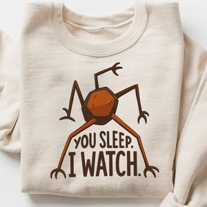May include: Cream-colored sweatshirt with a graphic of a brown insect-like creature. The text "YOU SLEEP. I WATCH." is printed below the creature. The sleeves are rolled up. A casual, comfortable garment.