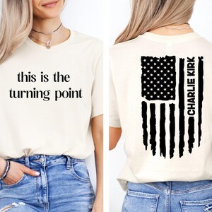 Peut inclure: T-shirt crème avec l'inscription "this is the turning point" sur le devant. Le dos du t-shirt présente un motif de drapeau américain noir et blanc avec le nom "CHARLIE KIRK" imprimé verticalement.