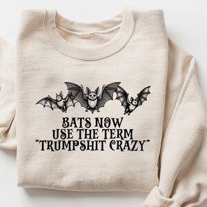 May include: Beige sweatshirt with a crew neck and long sleeves. The front features a graphic of three bats above the text: "BATS NOW USE THE TERM 'TRUMPSHIT CRAZY'" in black. The design is centered on the chest.