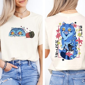 May include: Cream-colored t-shirt featuring a blue cartoon cat design on the front and back. The front design shows a cat peeking out with a potted plant. The back design has a larger cat with flowers and Japanese text.