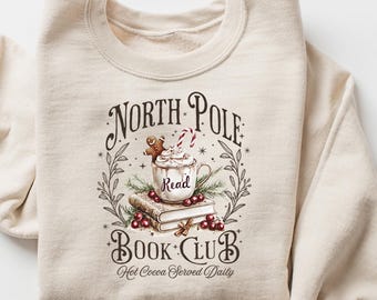 May include: Cream-colored sweatshirt featuring a graphic with the text "North Pole Book Club" and a mug of hot cocoa with a gingerbread man. The design includes books, berries, and greenery. The sleeves are rolled up.