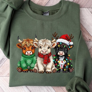 May include: An olive green sweatshirt with a graphic of three Highland cows. One cow is in a green sweater, another in a red scarf, and the third in a Santa hat and Christmas lights. The sweatshirt has long sleeves and a crew neck.