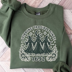 May include: An olive green sweatshirt with the text "Stirring Trouble Since 1692" in white. The graphic features three witch figures under a crescent moon and stars. The sweatshirt is folded on a light wood surface.
