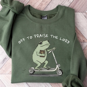 May include: An olive green sweatshirt with the phrase "Off to Praise the Lord" printed above a cartoon frog riding a scooter and holding a book. The sweatshirt has long sleeves and a crew neck.