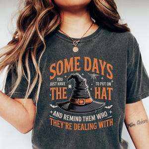 May include: Dark gray t-shirt with the text "SOME DAYS YOU JUST HAVE THE HAT TO PUT ON" in orange and white. The shirt features a witch hat graphic and is paired with blue jeans and white shoes. A Halloween-themed tee.
