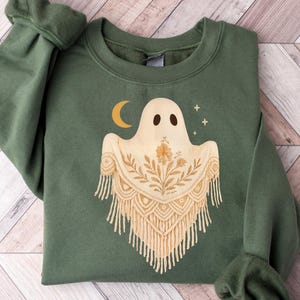 May include: Green crewneck sweatshirt featuring a beige ghost design with a crescent moon and stars. The ghost has a floral pattern and fringe detail. The sweatshirt is made of a soft material and is perfect for Halloween or everyday wear.